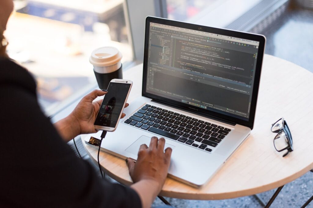 pexels photo 1181244 Developer working remotely, coding on a laptop with phone in hand, showcasing modern work culture.